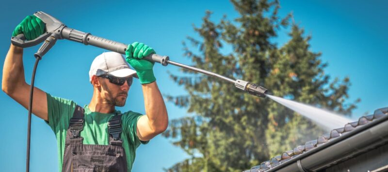 man wearing overalls holding a pressure washing machine while he pressure washes the roof of a house